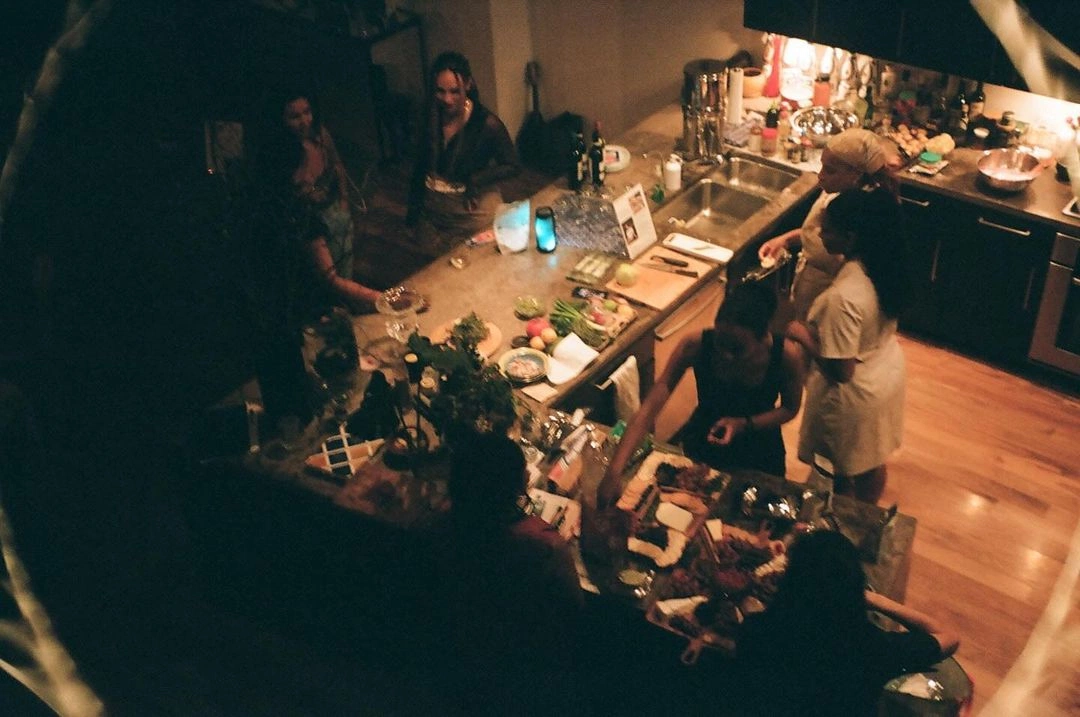 A warmly lit indoor evening gathering, people grouped around a kitchen.